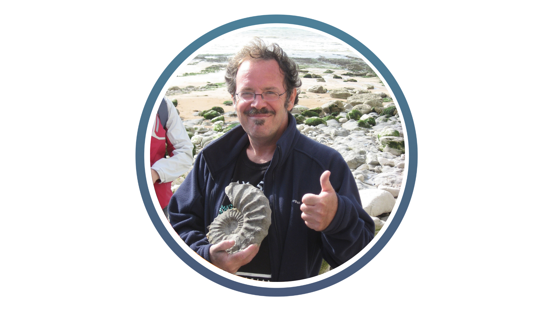male with glasses and moustache wearing a blue jacket and holding a large ammonite fossil