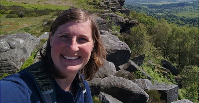 A woman with shoulder-length hair smiling at the camera with a rocky, hilly landscape and blue sky in the background