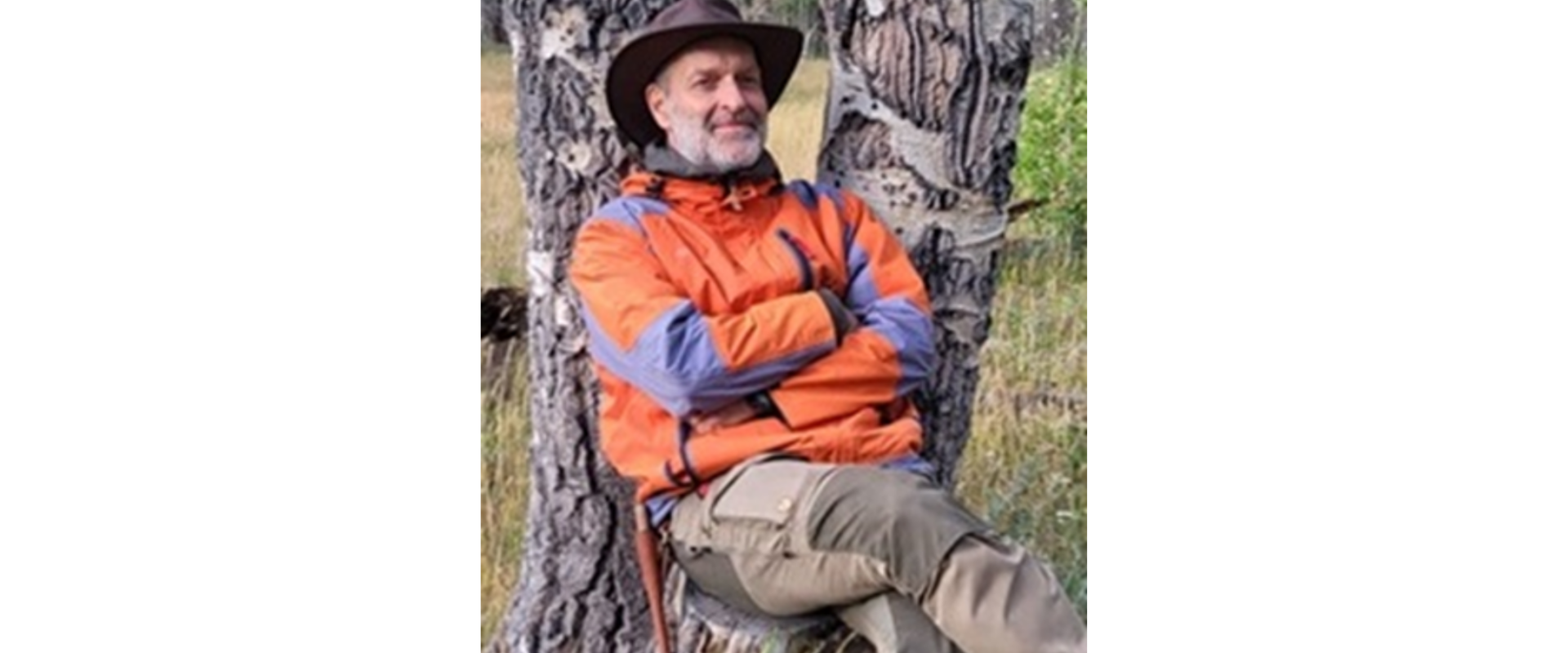 A person wearing a wide-brimmed dark hat, a bright orange and blue outdoor jacket, and khaki trousers reclines against a large fallen tree trunk in a wooded area. The tree bark is rough and partially charred or weathered. Grass and other trees are visible in the background. The image is rotated sideways, making the person appear horizontal across the frame.