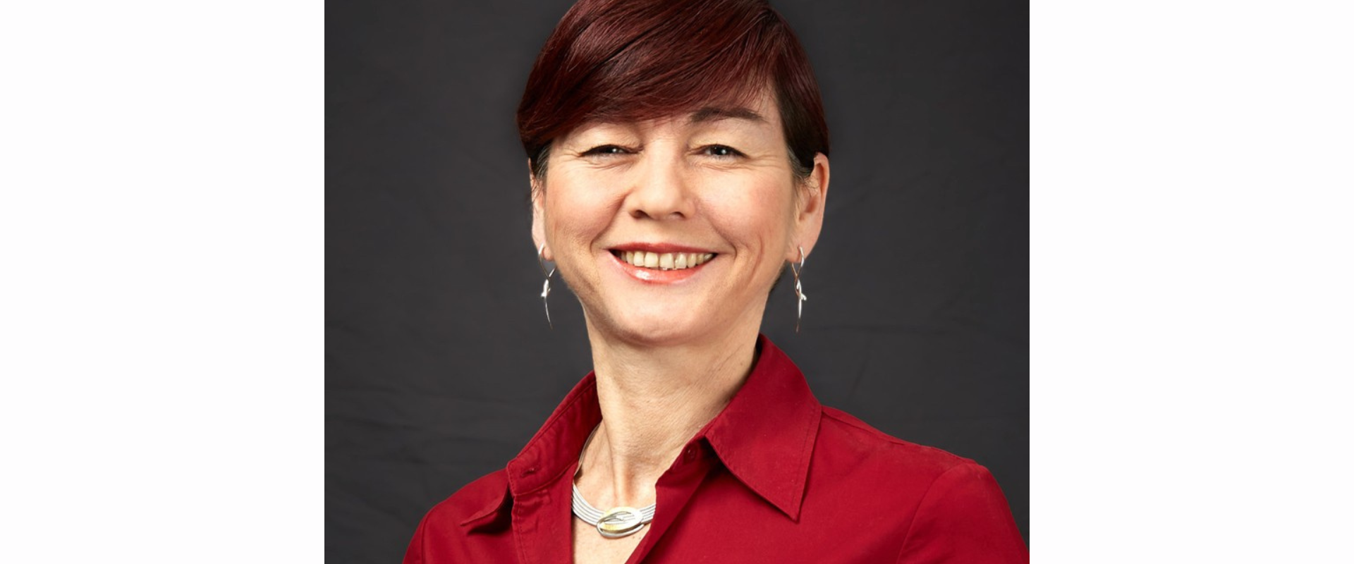 A person wearing a red collared shirt stands in front of a plain dark background. The person has short, neatly styled dark hair and is wearing a silver necklace and matching drop earrings.
