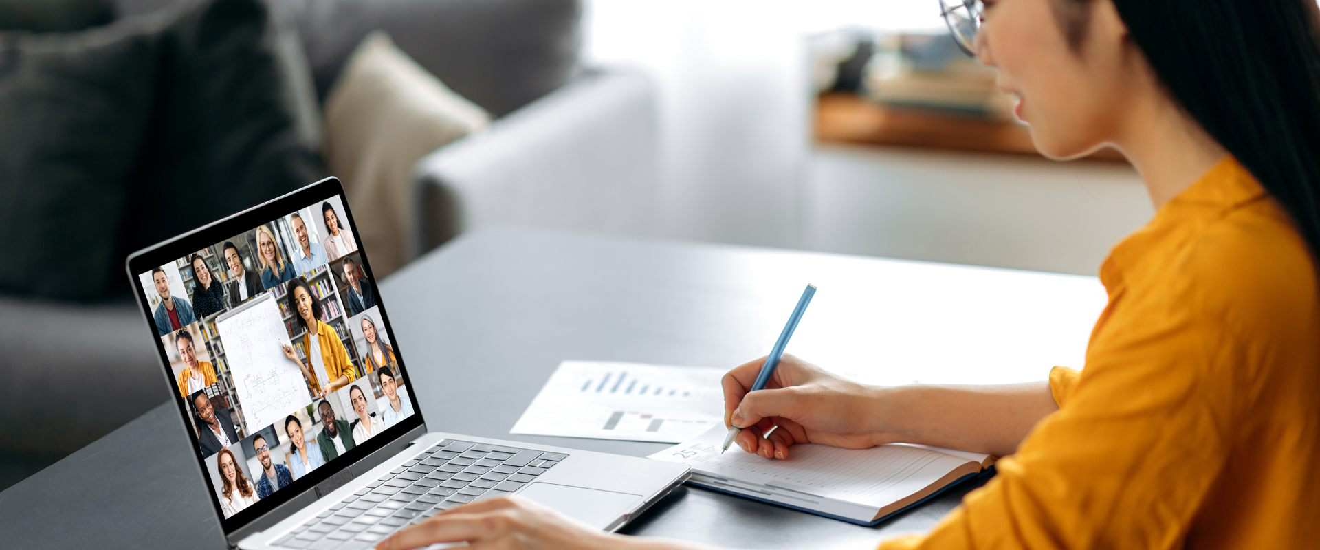 woman in a yellow jumper working at a laptop with many people on the screen on a video call