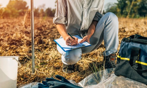 female crouching down in a field writing on a clipboard with research samples in the foreground