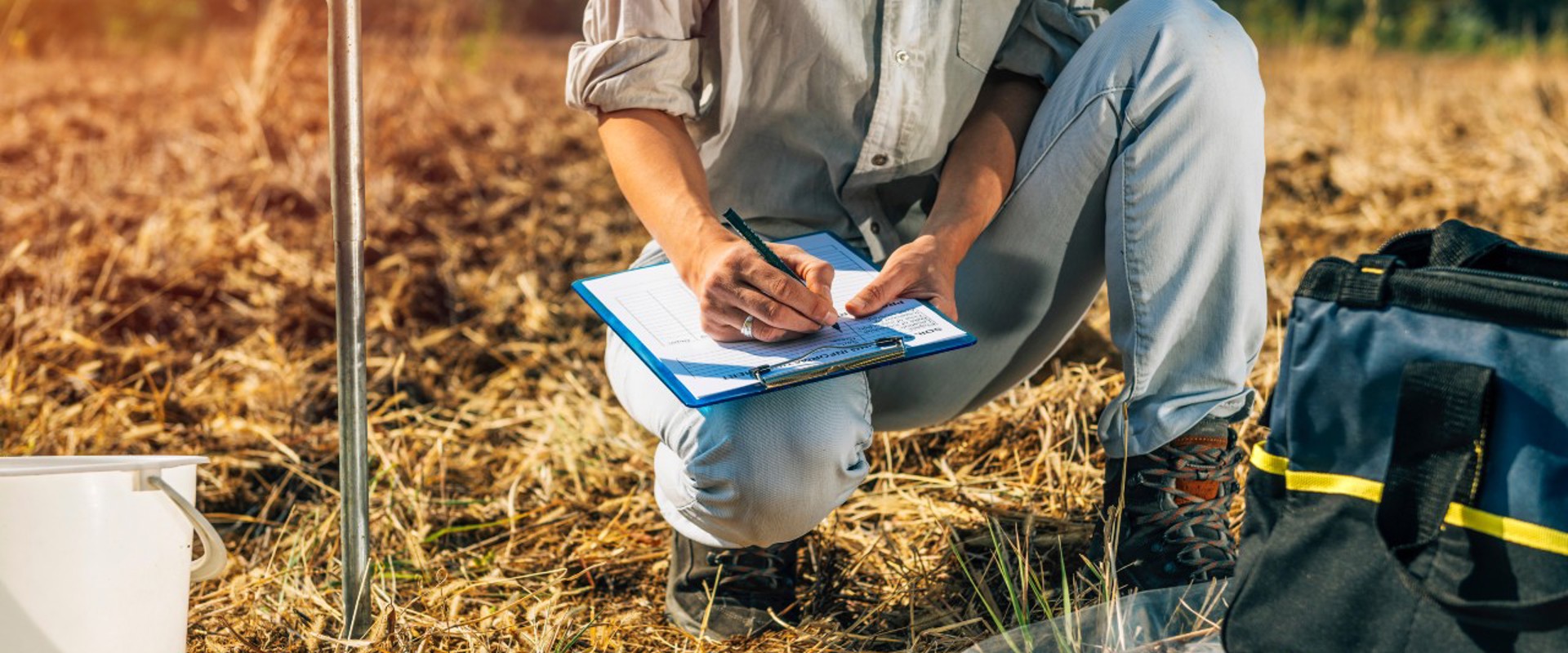 female crouching down in a field writing on a clipboard with research samples in the foreground