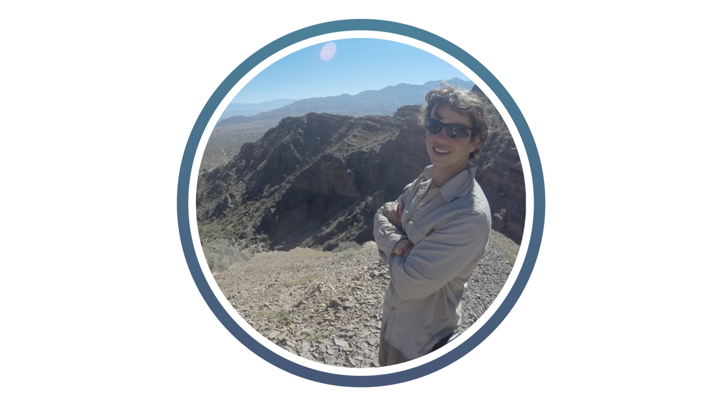 young white man with sunglasses on and a brown short stood in front of arid mountain landscape