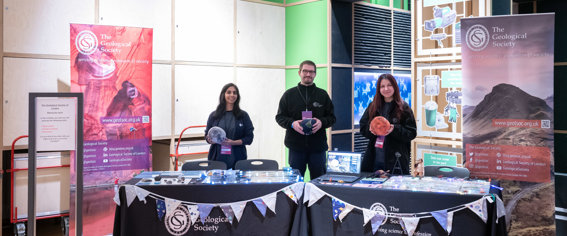 a white male, an Asian female and a white female standing at a table with various items on it and a geological society cloth covering it