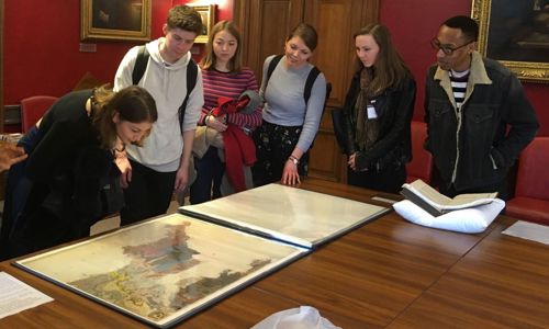 A group of six students in a grand room hung with portraits, looking at a geological map of Cornwall on a table