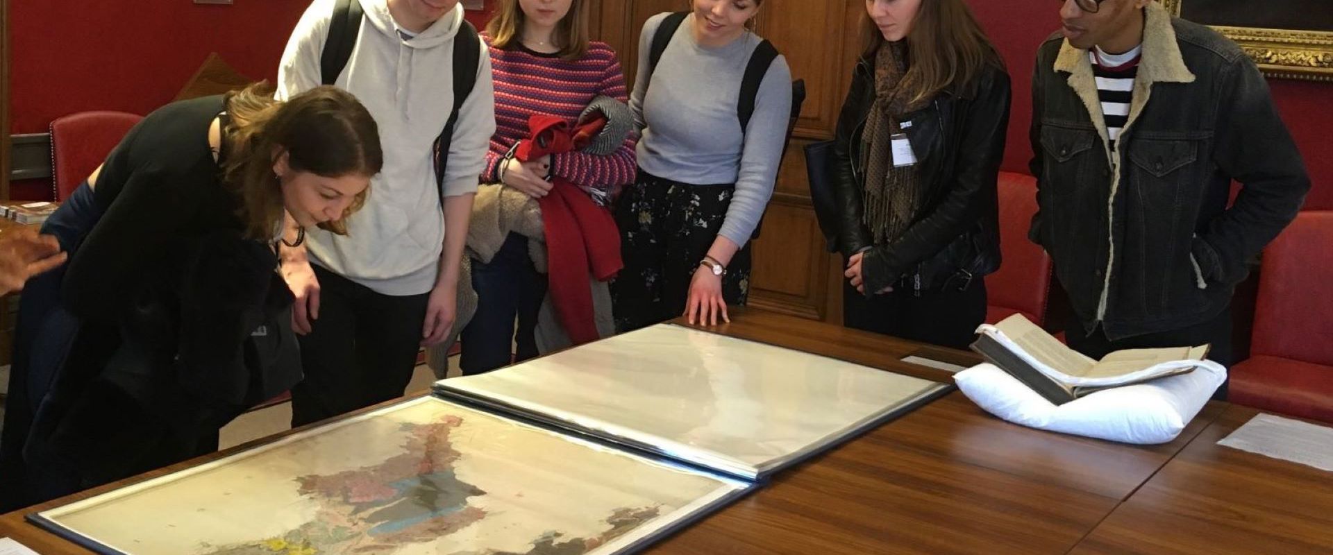 A group of six students in a grand room hung with portraits, looking at a geological map of Cornwall on a table