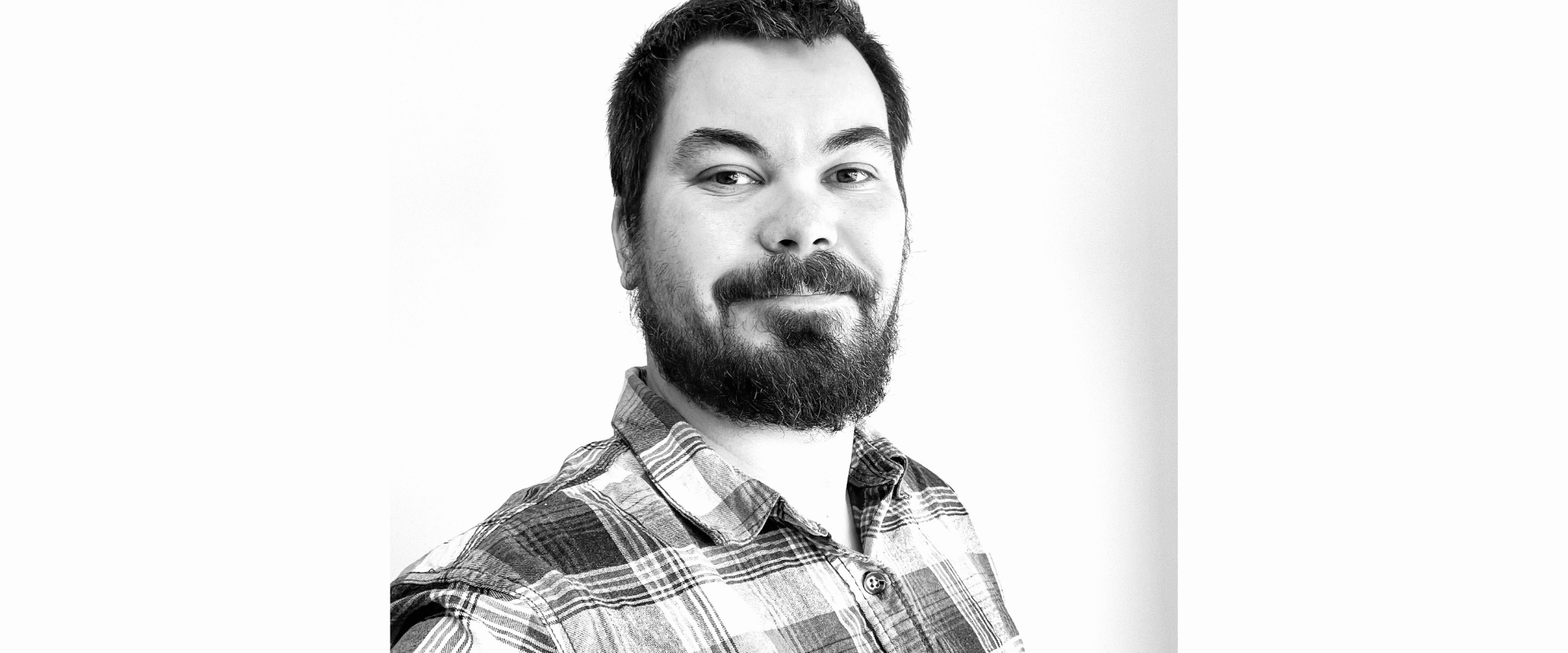 Black-and-white head-and-shoulders portrait of a person with short hair and facial hair, wearing a checkered button-up shirt. The person is positioned slightly to the left of centre and faces the camera at a slight angle. The background is plain and light-coloured, with soft, even lighting and no visible text or objects.