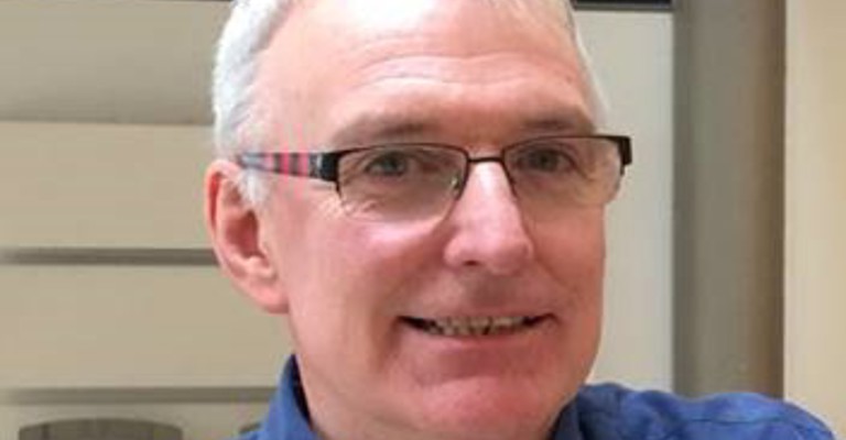 A man with short white hair wearing glasses and a blue shirt smiling at the camera