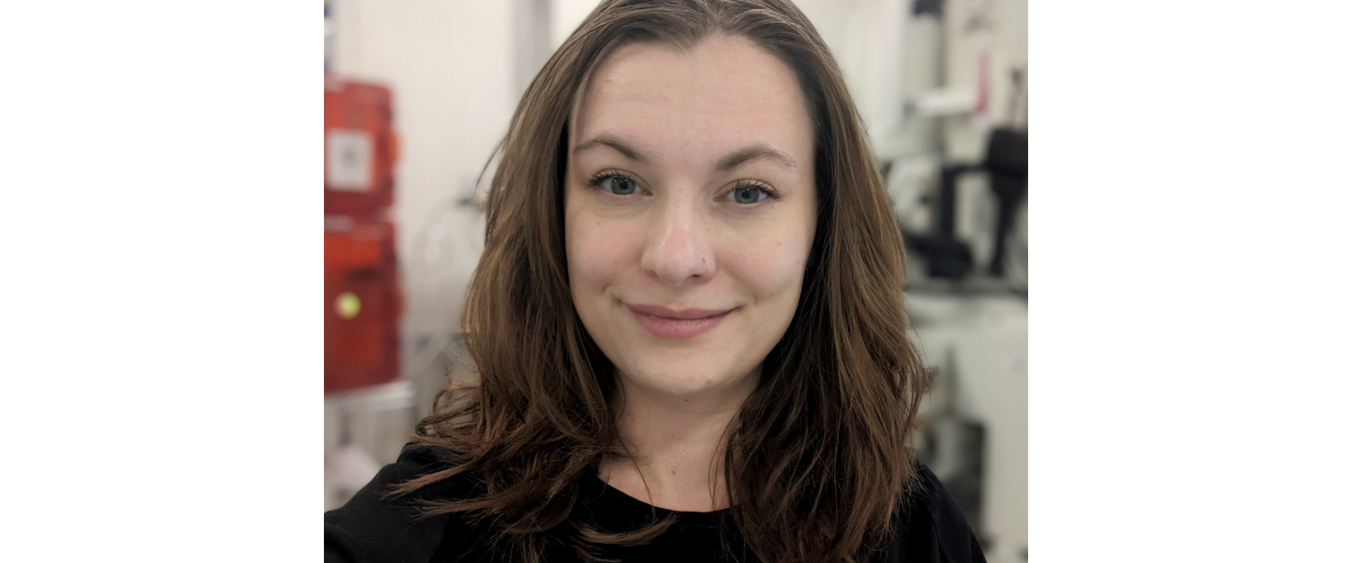 Close-up portrait of a person with shoulder-length brown hair wearing a black top, facing the camera indoors. The background is softly blurred, showing a bright workspace or laboratory setting with equipment and a red container visible behind the person. Lighting is even and natural, with the person centered in the frame.
