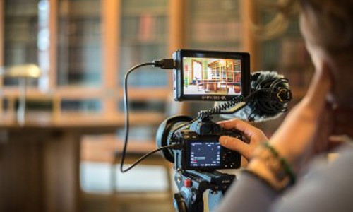 A person recording a video in a library, surrounded by books and shelves, focused on their camera.