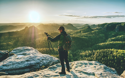 man in outdoor style clothing holding a tripod and stood atop a mountain 