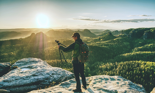 man in outdoor style clothing holding a tripod and stood atop a mountain 