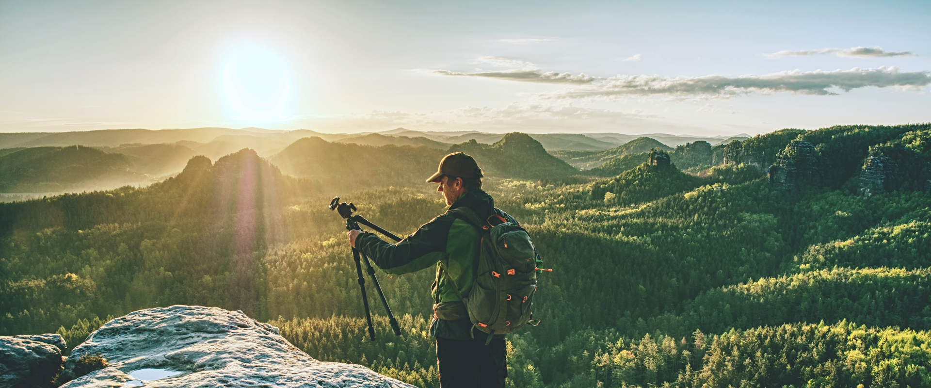 man in outdoor style clothing holding a tripod and stood atop a mountain 