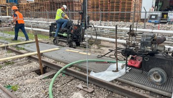 Two construction workers wearing safety gear operate drilling equipment along a set of railway tracks. One worker is seated on a small drilling rig, while another stands nearby. The scene includes hoses, machinery, and stacks of wooden pallets in the background.