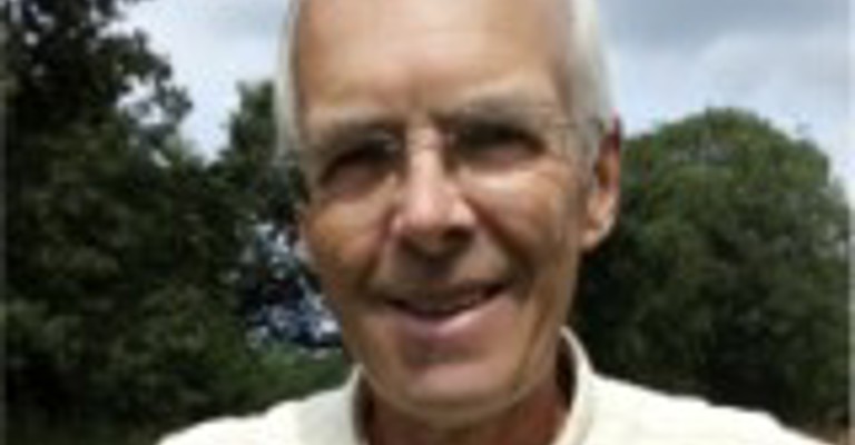 A man with wearing glasses and a pale yellow shirt smiling at the camera