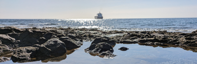 Looking out to the sea from a rocky shore, with a blue sky in the background and a boat on the horizon