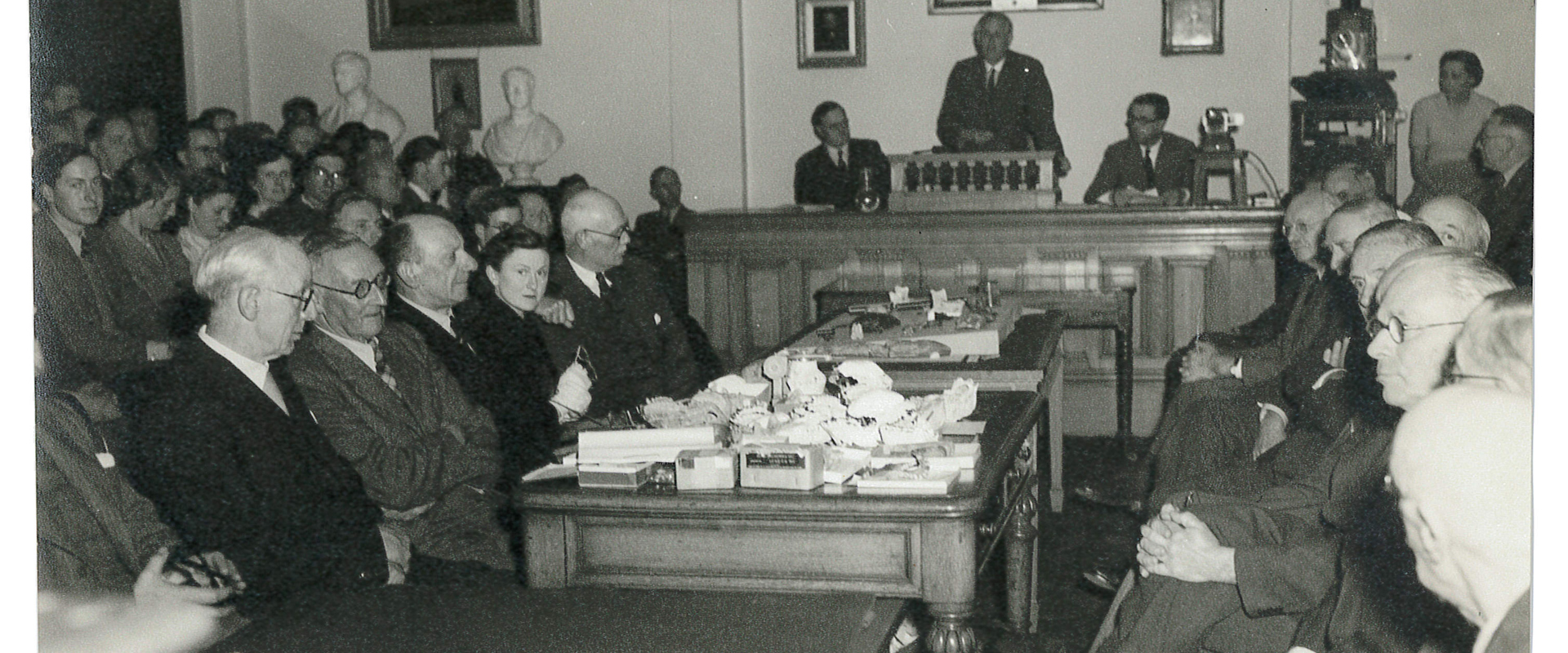 Old photograph of individuals seeting in a room facing each other durint a meeting