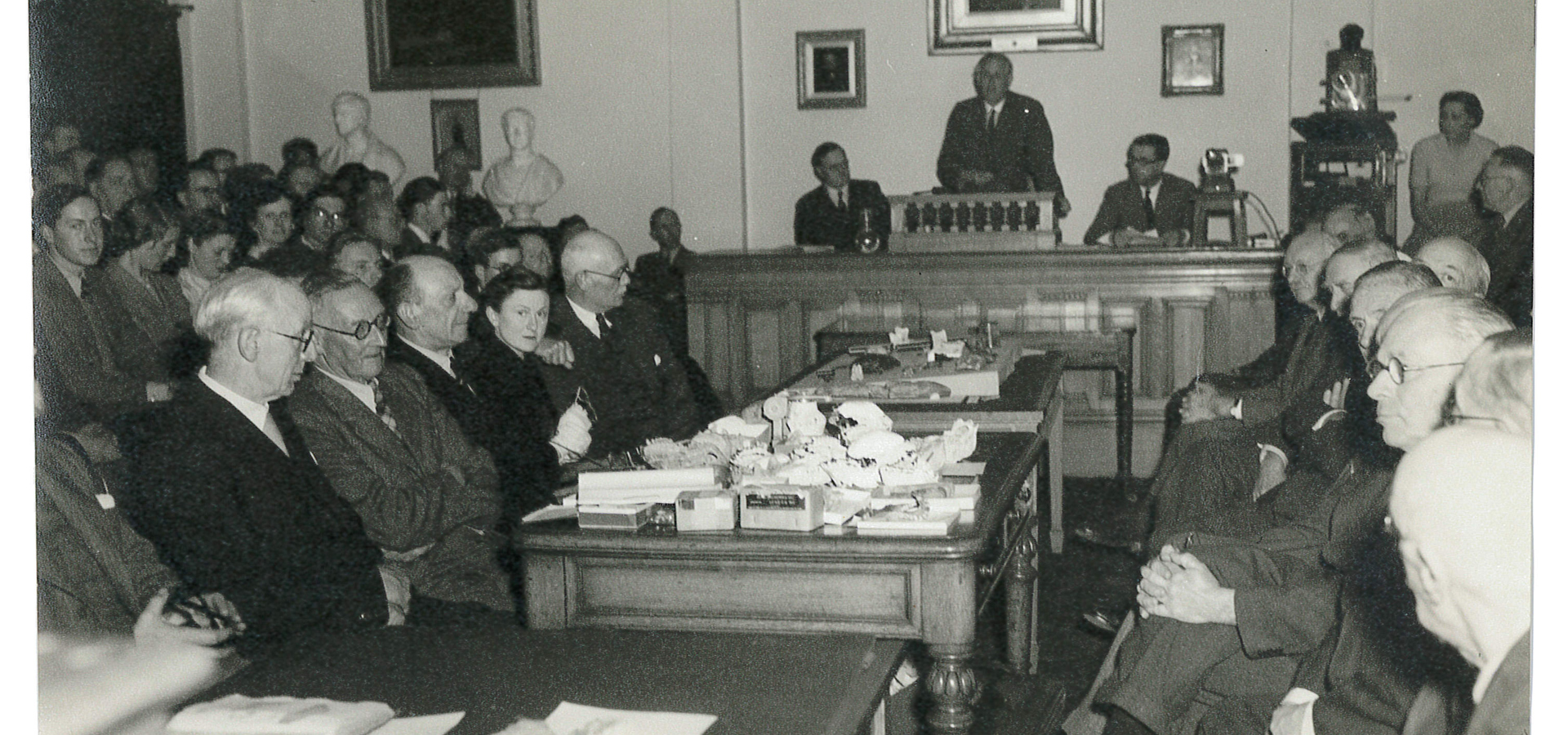 Old photograph of individuals seeting in a room facing each other durint a meeting