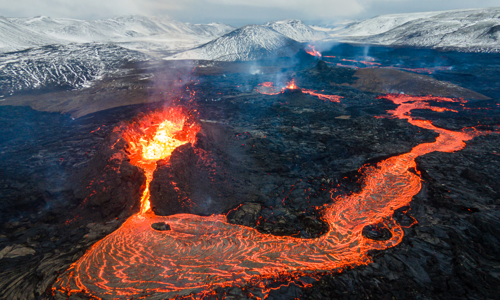 aerial view of a volcano spilling magma out, with the magma flowing like a river down the mountainside and snow covered mountains in the background