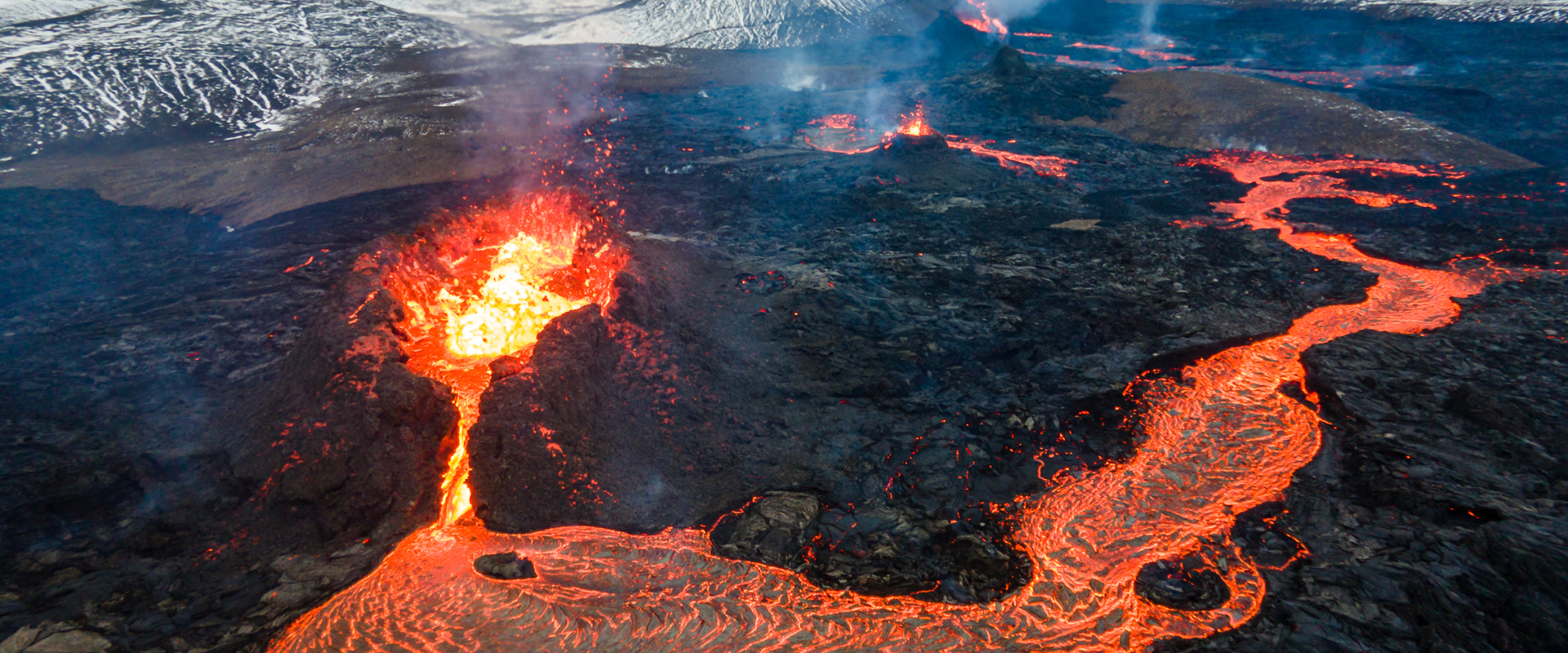 aerial view of a volcano spilling magma out, with the magma flowing like a river down the mountainside and snow covered mountains in the background