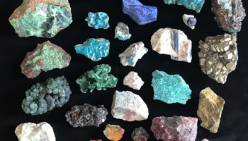 A collection of colourful mineral and rock specimens arranged in a grid on a black background. The assortment includes pieces in shades of turquoise, green, blue, purple, white, gold, and brown, with varied textures such as crystalline surfaces, rough edges, metallic clusters, and smooth formations.