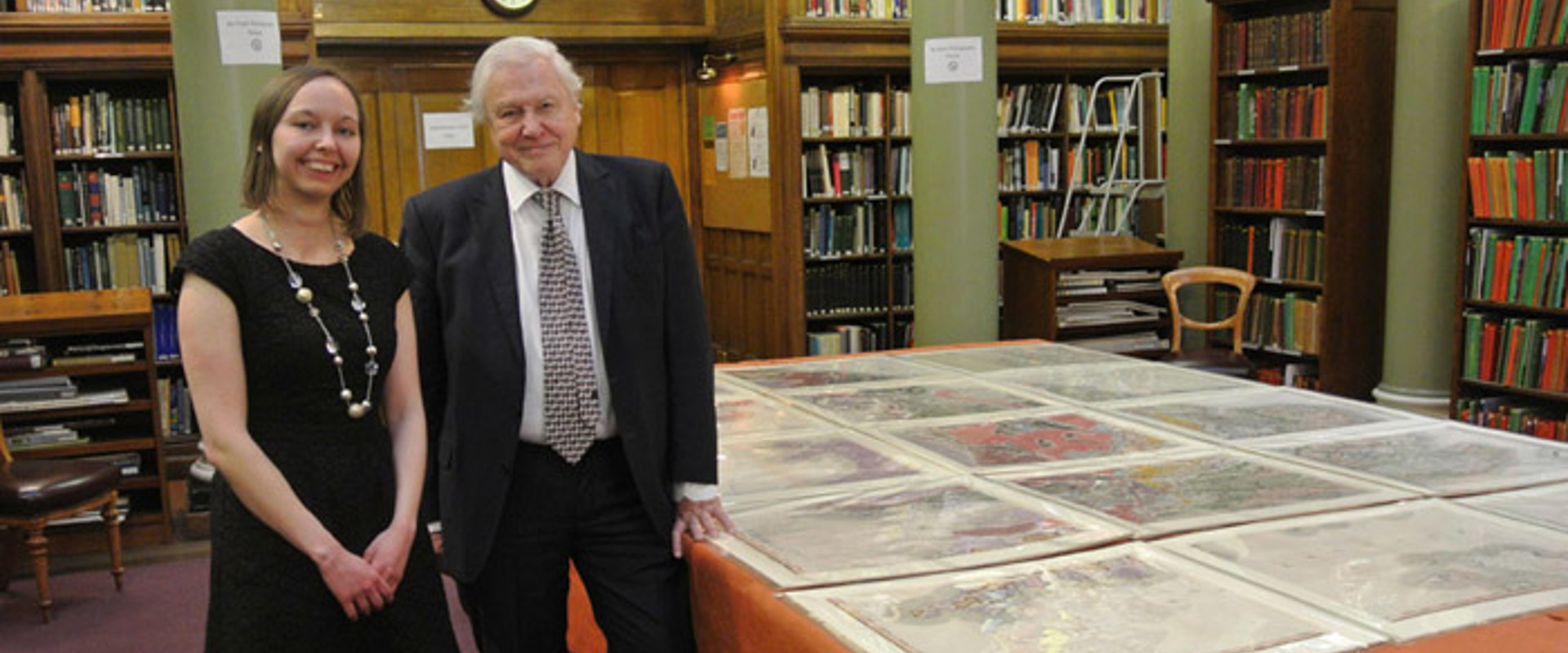 Victoria Woodcock and Sir David Attenborough standing in front of William Smith's map in the Geological Society's Upper Library