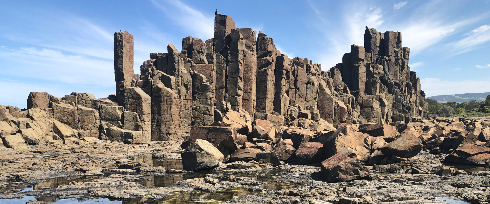 Dramatic rock formations rise against a bright blue sky, showcasing tall, angular columns. The foreground features rugged, rocky terrain.
