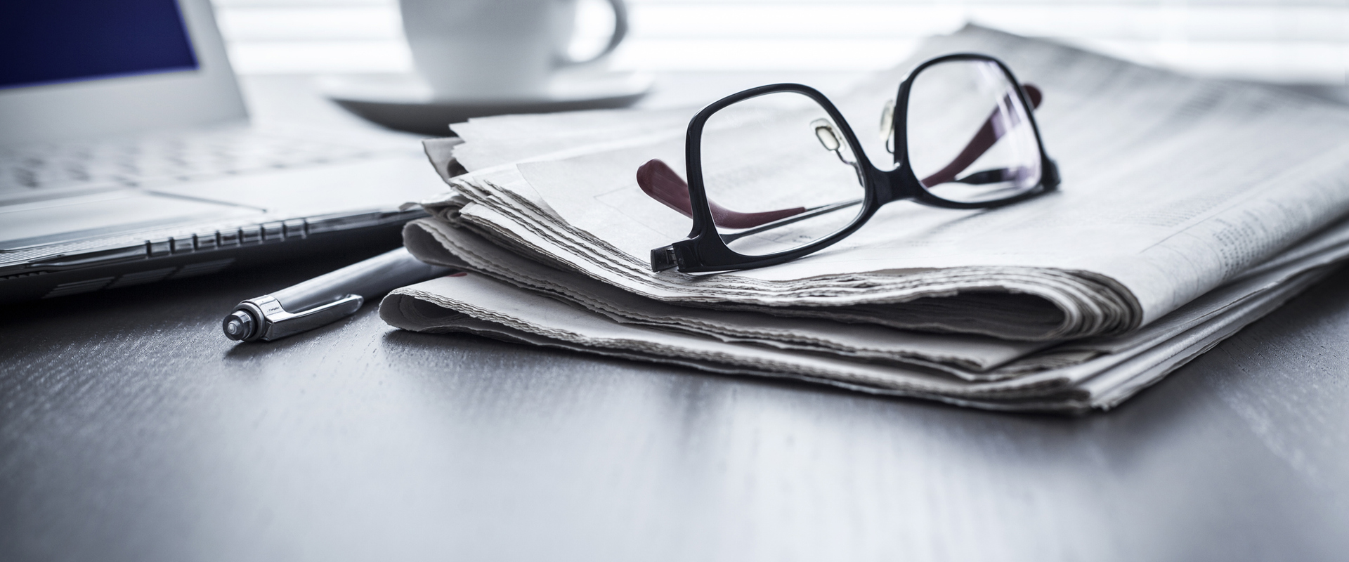 newspapers on a desk along with a pair of glasses, a pen, a cup of coffee and an open lap top