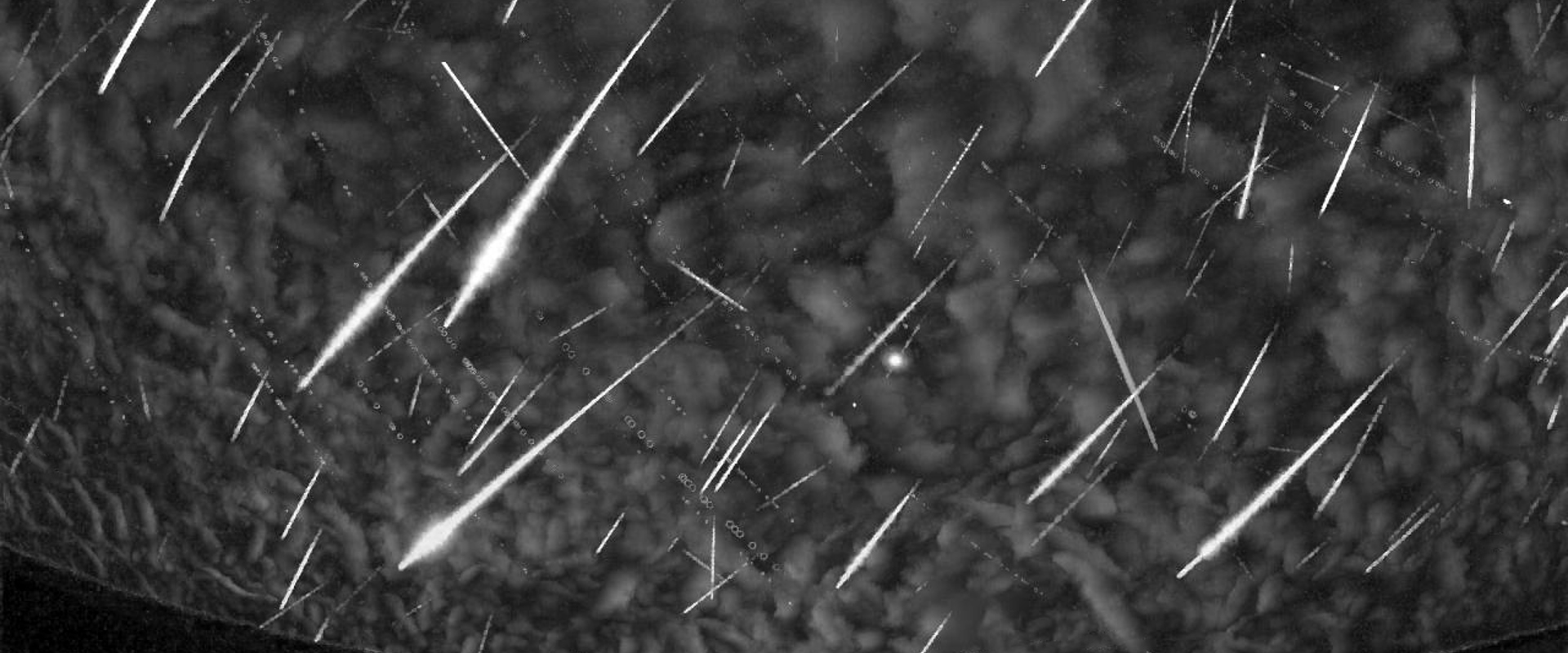 A black‑and‑white image showing numerous bright, thin, elongated streaks and lines scattered across a textured, grainy background. The streaks vary in length and angle, creating a dynamic, radiating pattern. A small circular marker is visible near the centre of the image.