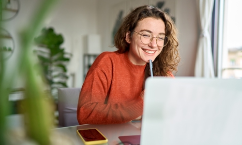Young woman in glasses smiling at laptop with pen