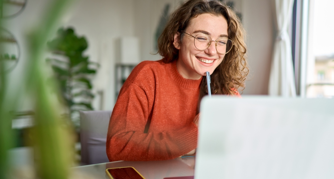 Young woman in glasses smiling at laptop with pen