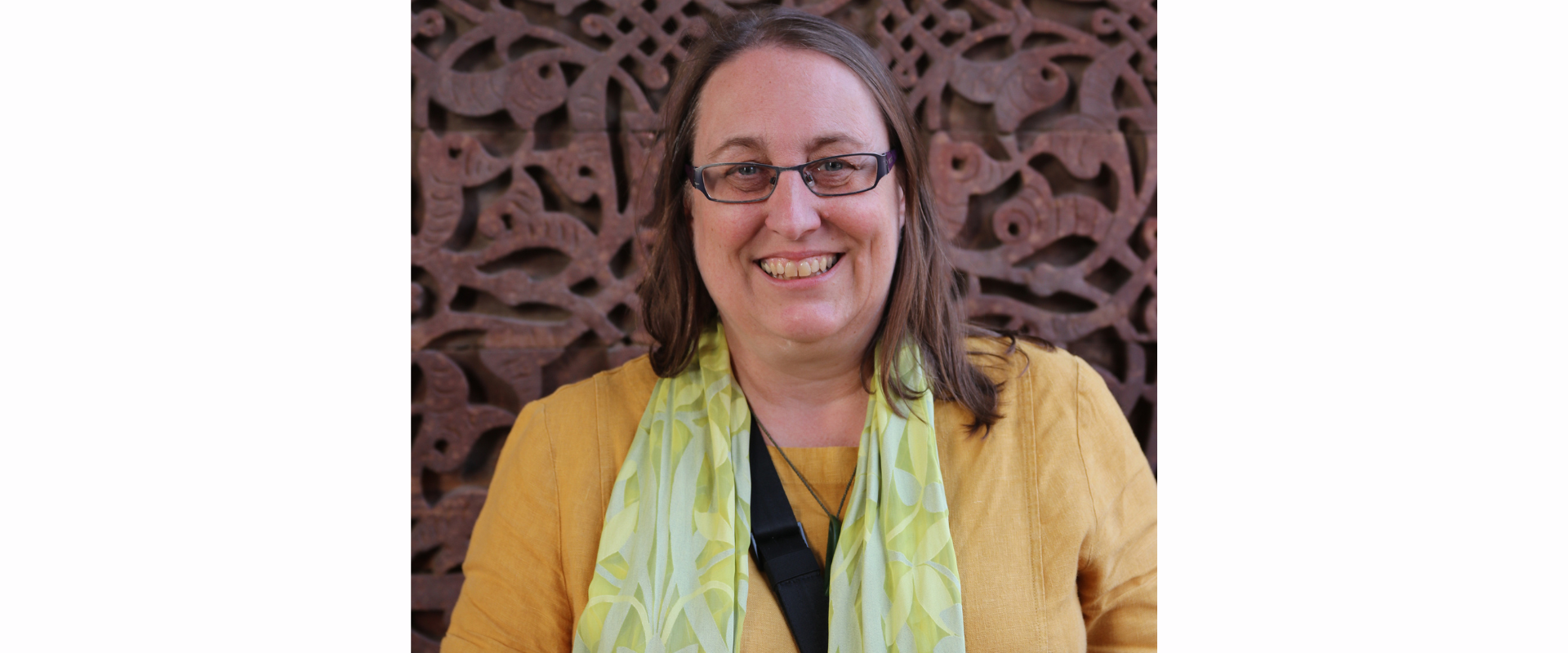 A person wearing a mustard‑coloured top with a light green patterned scarf, standing in front of an ornate carved backdrop featuring intricate swirling shapes and cut‑out details. A black strap is visible across the person’s chest.