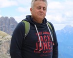 A man wearing a dark hoodie and backpack stands outdoors with mountains and a partly cloudy sky in the background.
