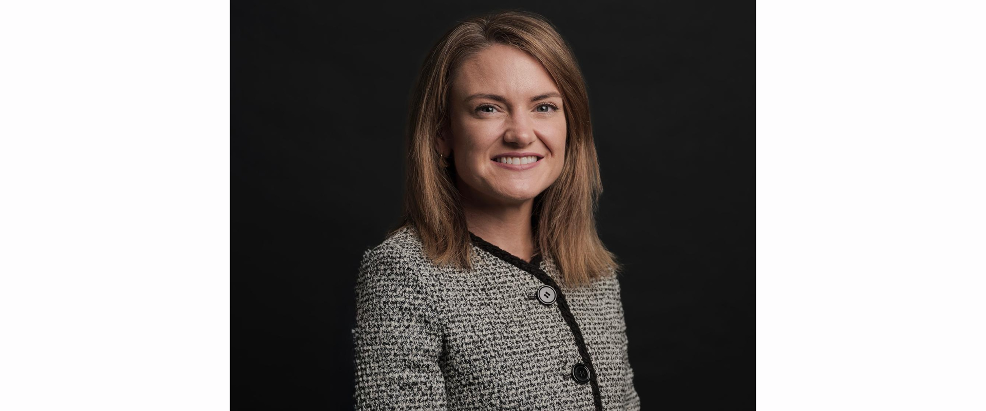 A person wearing a textured, light‑coloured jacket with dark trim and large buttons, posed against a solid black background. The person has shoulder‑length hair and is positioned slightly angled to one side.