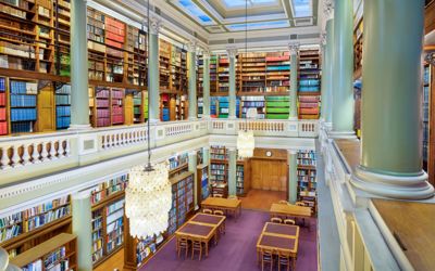 The Upper Library of the Geological Society of London, taken from an upstairs gallery looking down at table and chairs on a maroon carpet surrounded by book-lined shelves. Two bright glass chandeliers hang above the library.