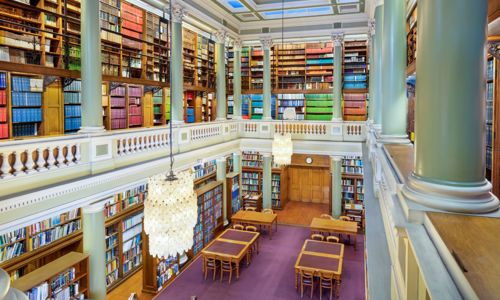 The Upper Library of the Geological Society of London, taken from an upstairs gallery looking down at table and chairs on a maroon carpet surrounded by book-lined shelves. Two bright glass chandeliers hang above the library.