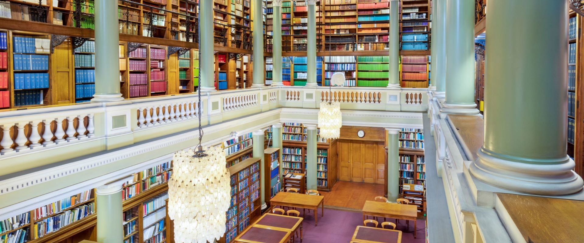 The Upper Library of the Geological Society of London, taken from an upstairs gallery looking down at table and chairs on a maroon carpet surrounded by book-lined shelves. Two bright glass chandeliers hang above the library.