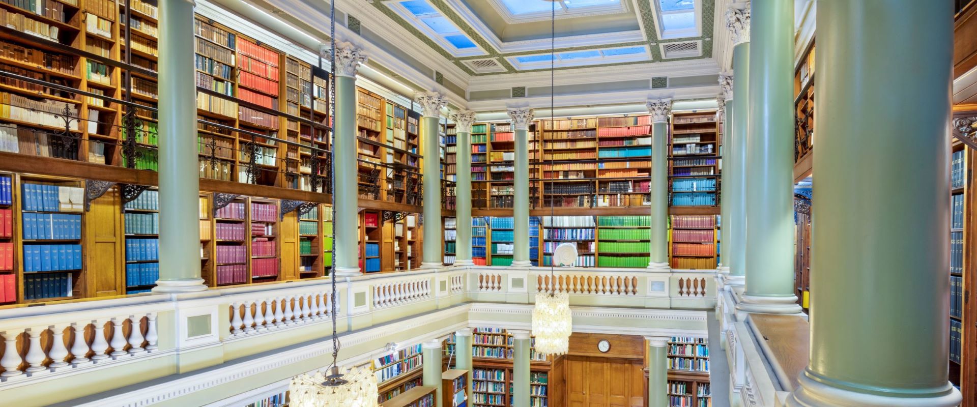 The Upper Library of the Geological Society of London, taken from an upstairs gallery looking down at table and chairs on a maroon carpet surrounded by book-lined shelves. Two bright glass chandeliers hang above the library.
