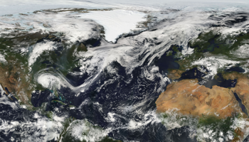 Satellite view of Earth showing North America, Greenland, Europe, and Africa with swirling white clouds. Ice covers Greenland; deserts are visible in Africa.