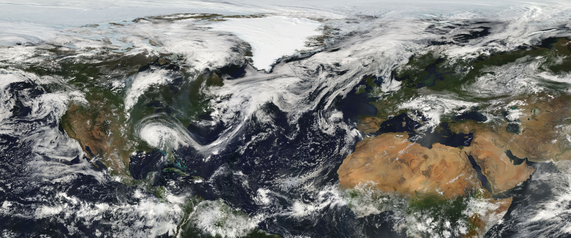 Satellite view of Earth showing North America, Greenland, Europe, and Africa with swirling white clouds. Ice covers Greenland; deserts are visible in Africa.