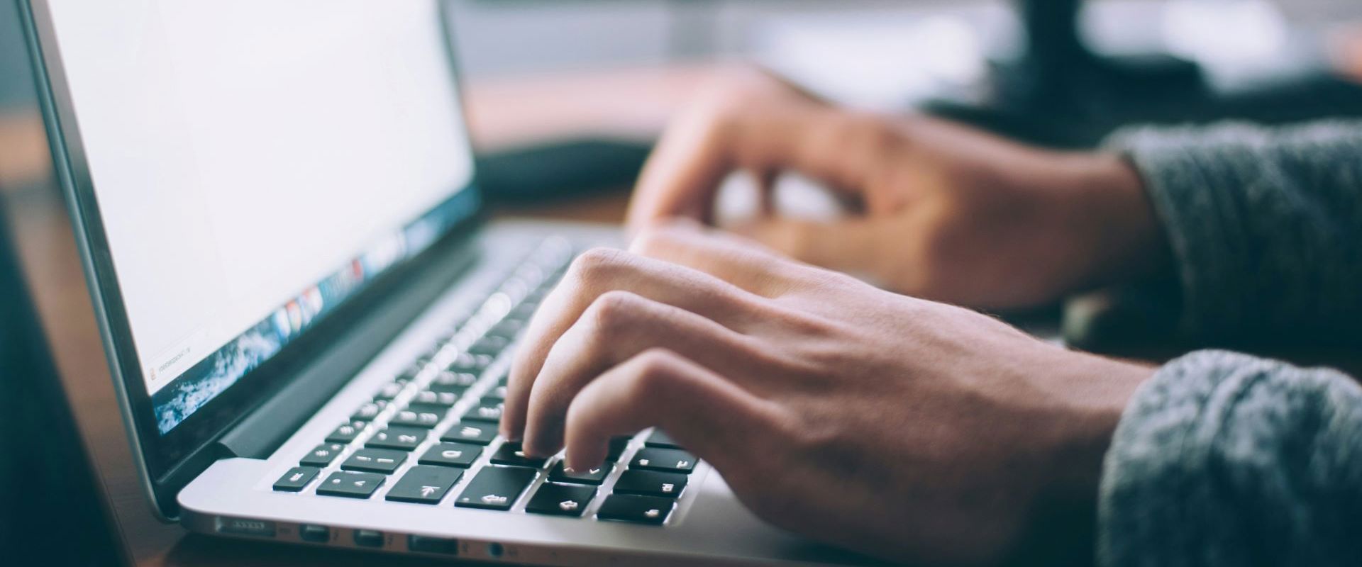 A laptop with two hands typing on it, with a PC blurred in the background