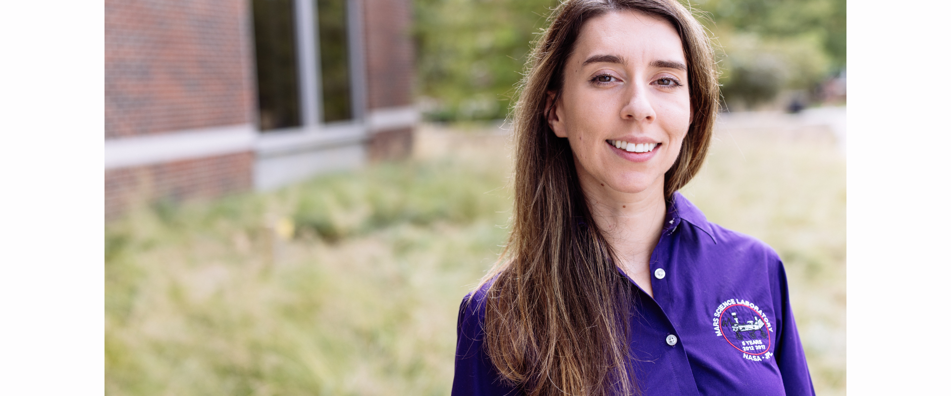 A person wearing a purple collared shirt with an embroidered circular logo on the left side of the chest, standing outdoors near a building with large windows and surrounded by grass and greenery.