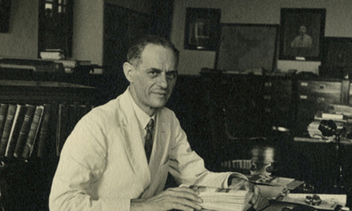 black and white image of an older white man wearing a white coat with a shirt and tie underneath. The man is sitting a a desk and has an open book in his hands