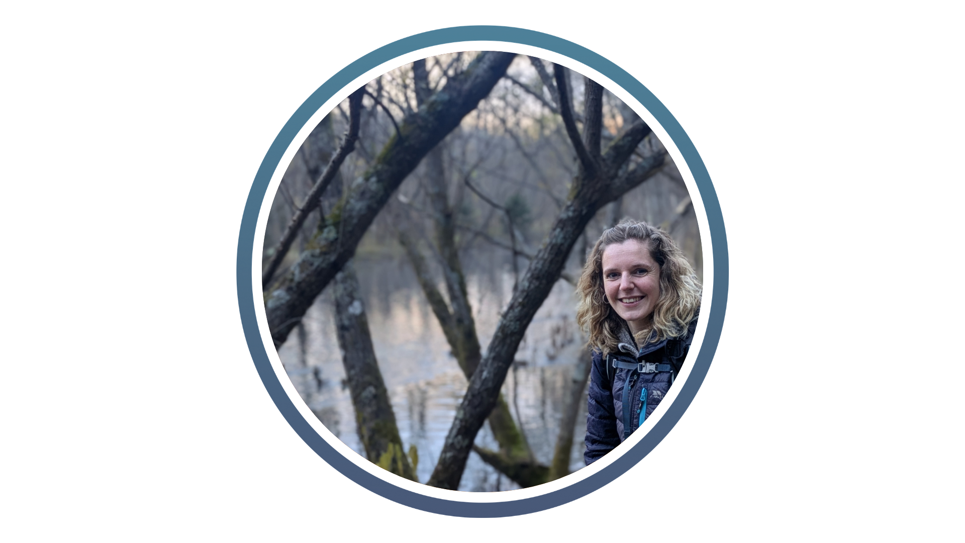 A woman with curly hair smiles by a lakeside. Bare trees frame the scene, and the water reflects the overcast sky, creating a serene atmosphere.