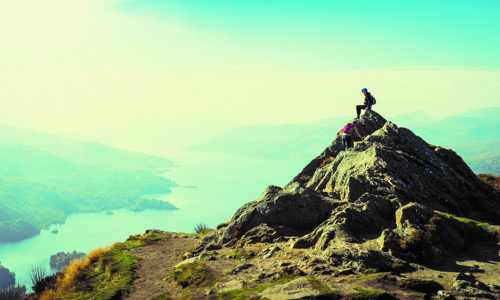 Two people in hard hats climb a pointed rock on a cliff above a stretch of lake