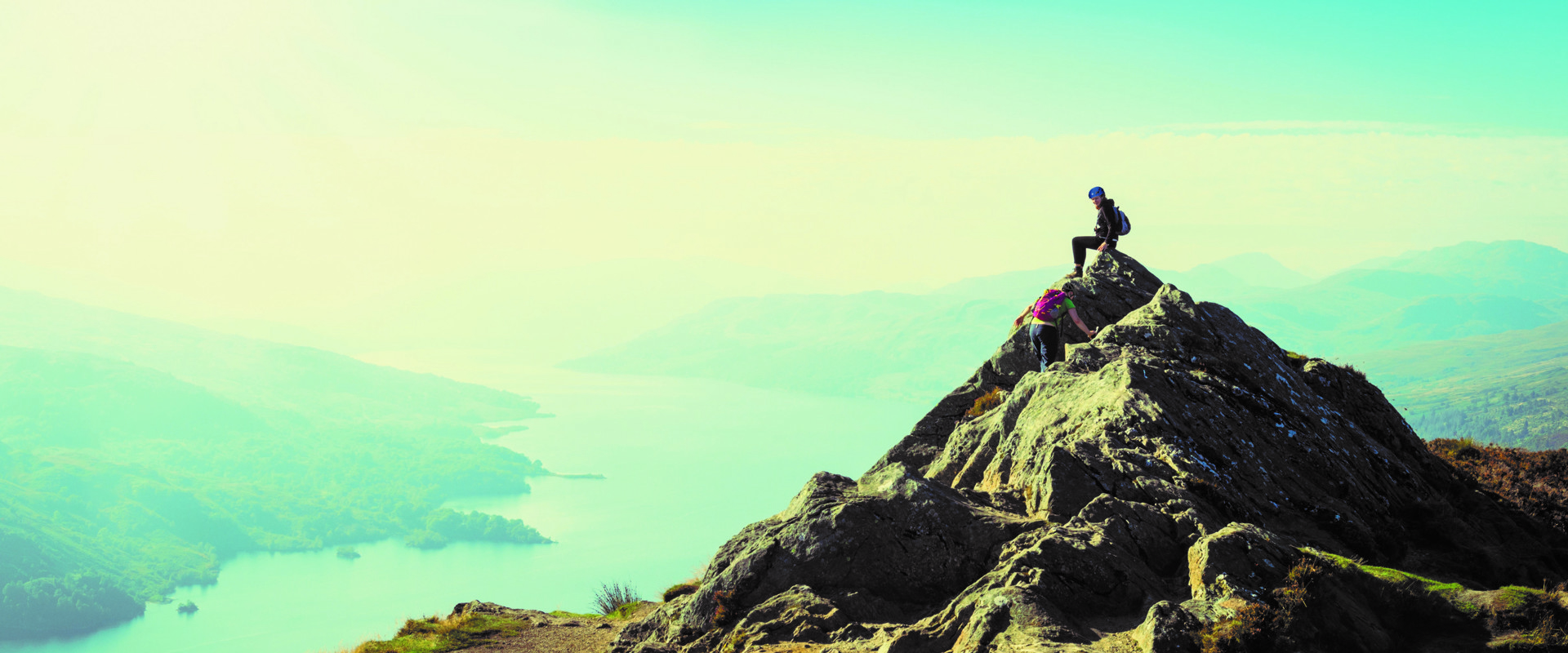 Two people in hard hats climb a pointed rock on a cliff above a stretch of lake
