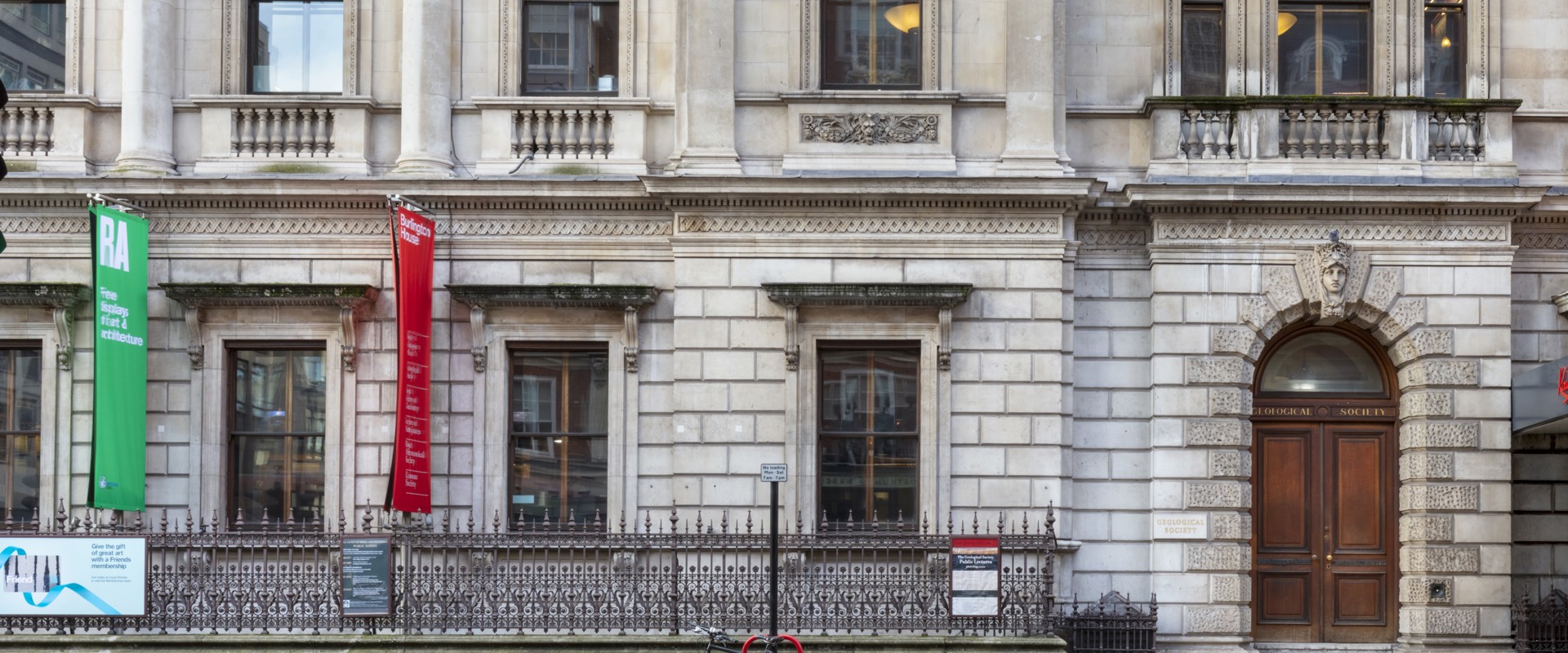 Facade of an ornate stone building with tall windows, decorative molding, and banners hanging on the left side. A metal fence runs along the front.