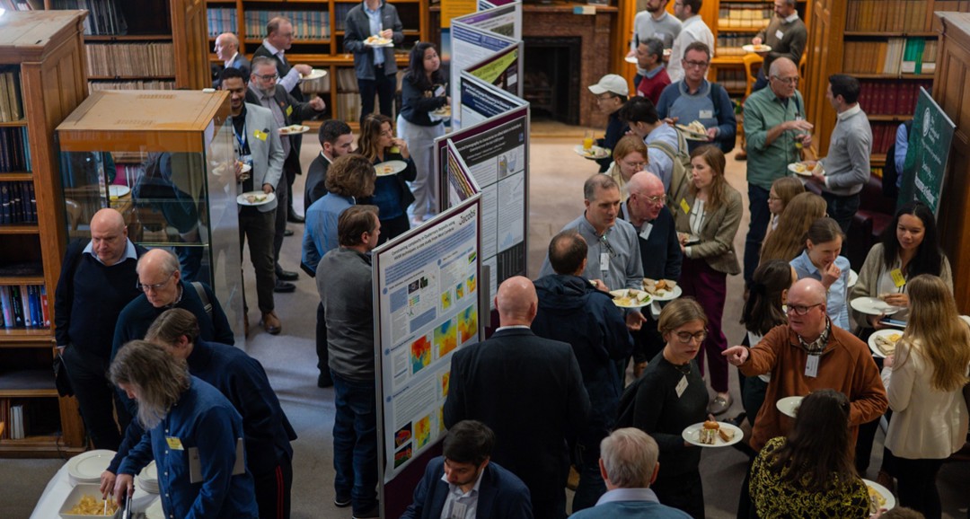 A lively networking event in a library, with attendees enjoying food and discussing onlookers among colorful posters and bookshelves.