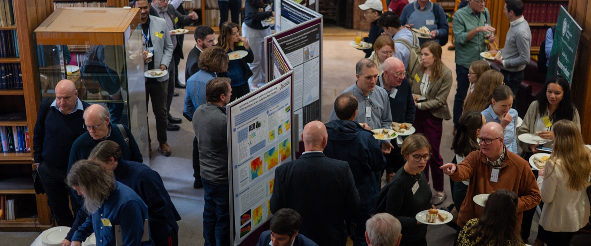 A lively networking event in a library, with attendees enjoying food and discussing onlookers among colorful posters and bookshelves.
